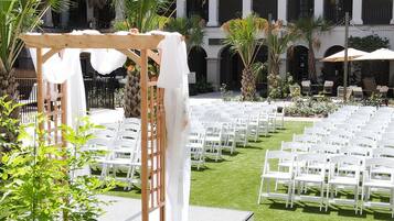 An outdoor wedding venue with white chairs arranged in rows, a wooden archway, and a building with columns in the background.