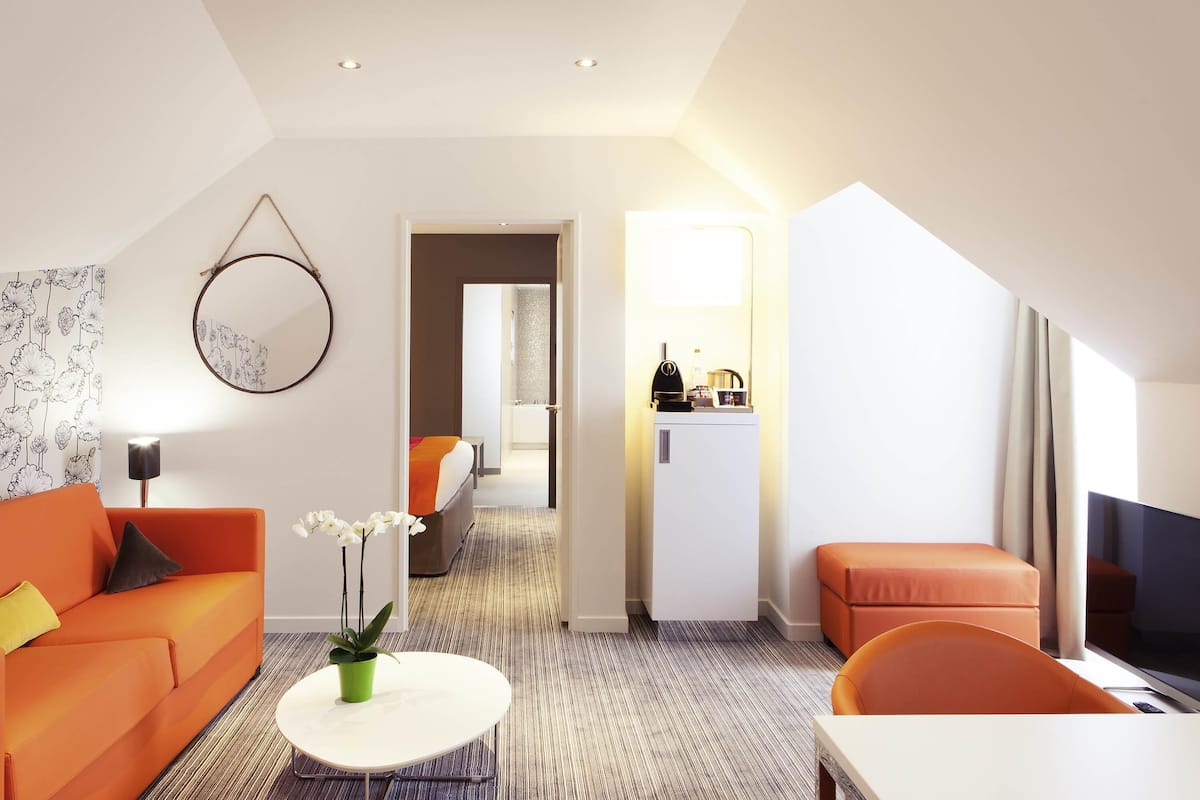 A modern hotel room with a sofa, a small table with a plant, a bed with an orange blanket, a TV, and a kitchenette.