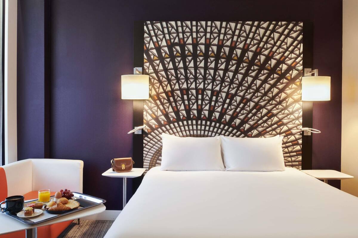 Una habitación de hotel con cama, una mesa con desayuno, dos lámparas de noche y un papel tapiz estampado.