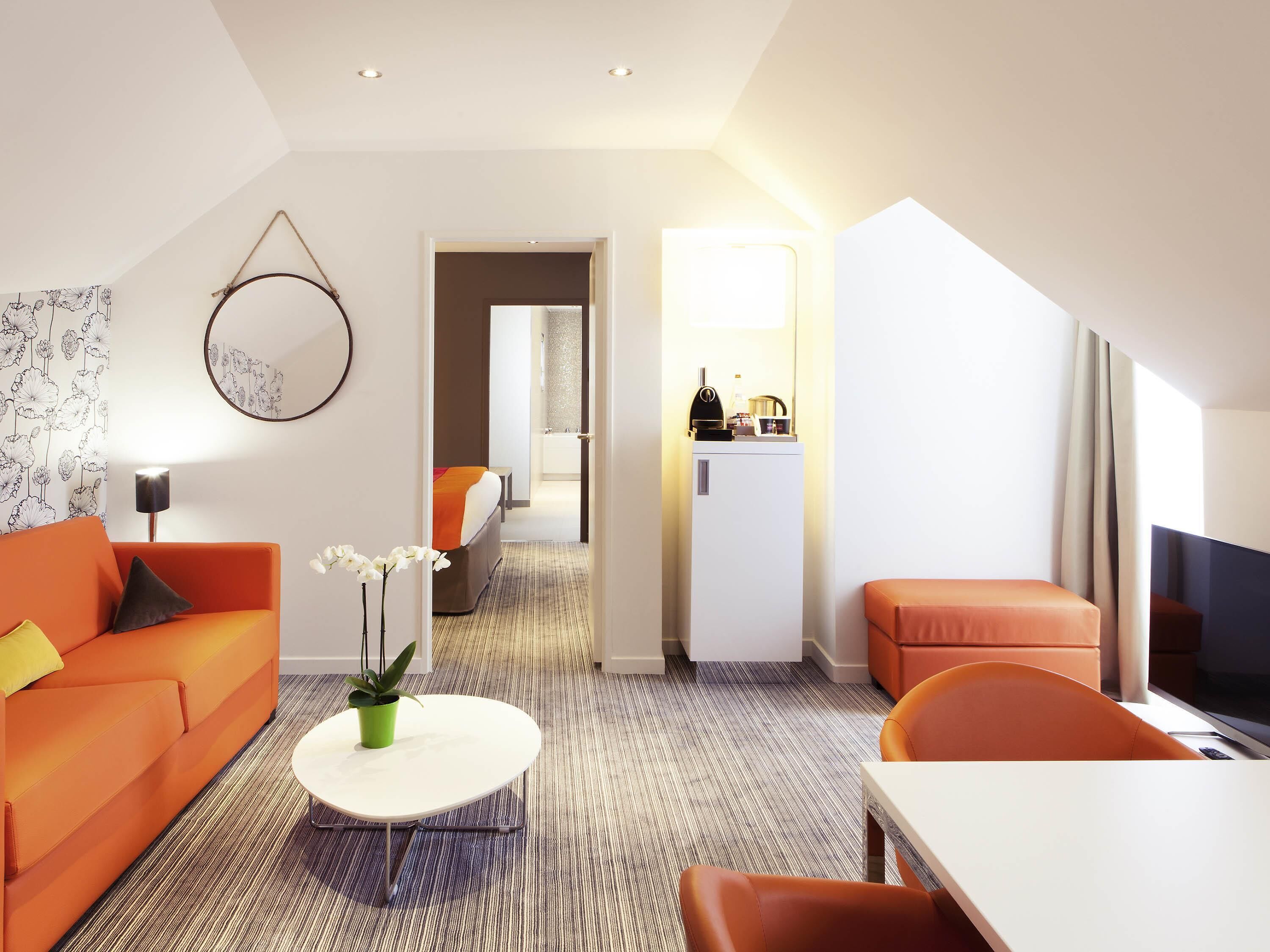 Una habitación de hotel moderna con un sofá, una mesita con una planta, una cama con una colcha naranja, un televisor y una kitchenette.