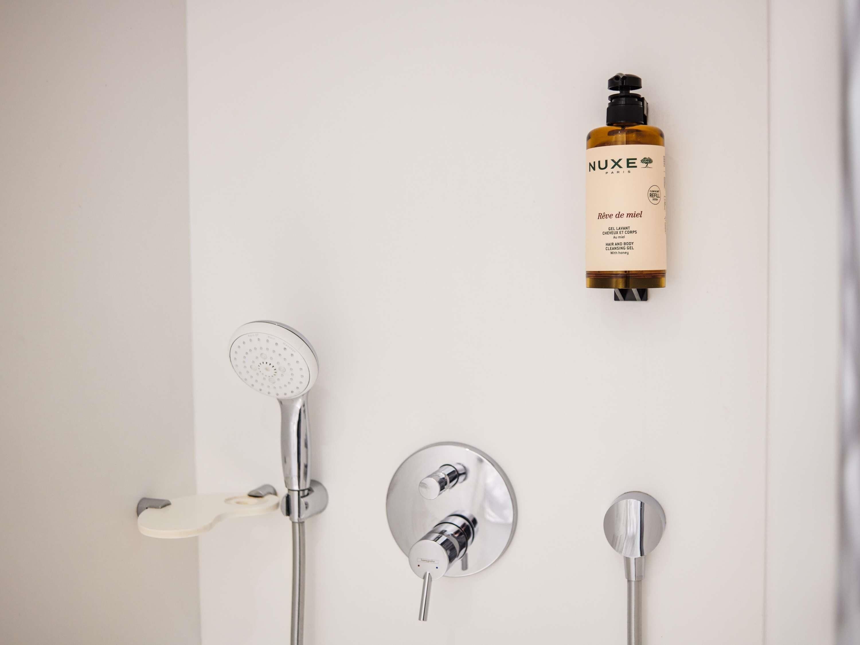 A bathroom with a shower, a handheld showerhead, and a bottle of NUXE Paris body wash.