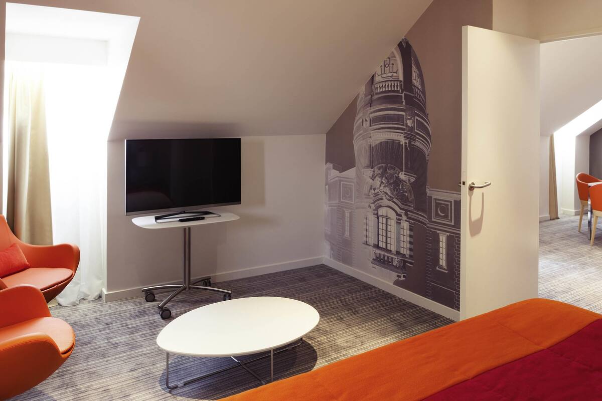 Habitación de hotel con televisor de pantalla plana, una pequeña mesa, un sofá naranja y un mural de un edificio en la pared.