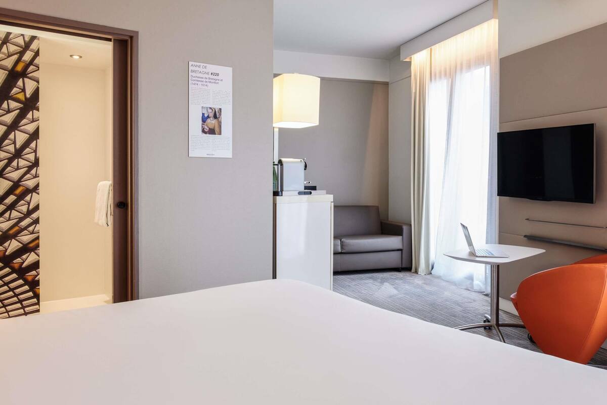 Una habitación de hotel con cama, escritorio con computadora portátil, televisión, silla y un cartel en la pared.