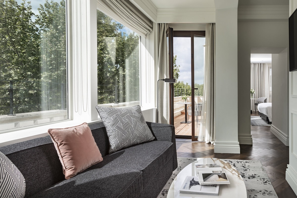 Una sala de estar moderna con un sofá gris, una mesa de centro con revistas y un ventanal que ofrece vistas a árboles y un balcón.