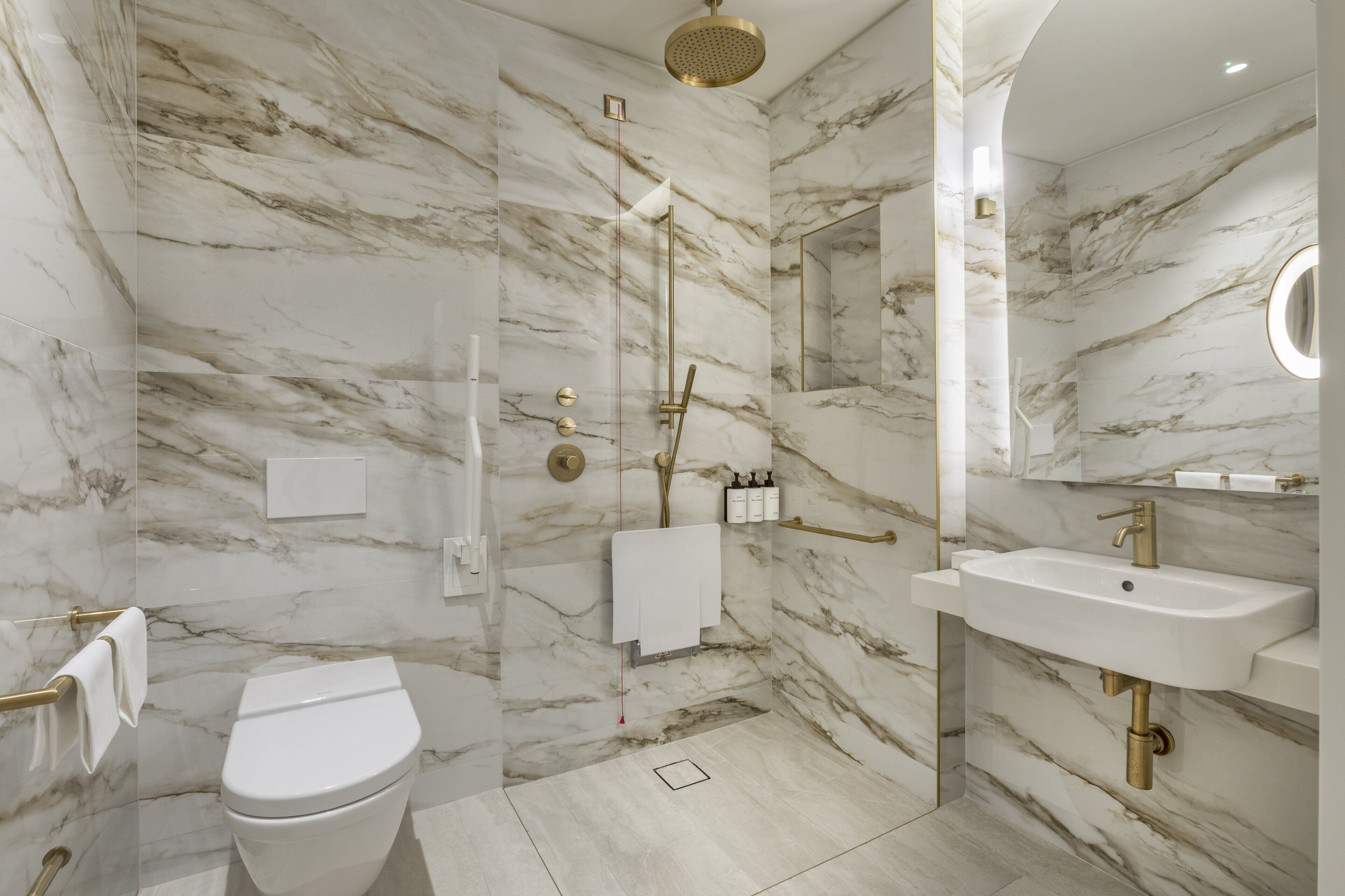 Une salle de bain moderne avec des murs en marbre, des toilettes, une douche dotée d’un pommeau doré et un lavabo avec un robinet doré.