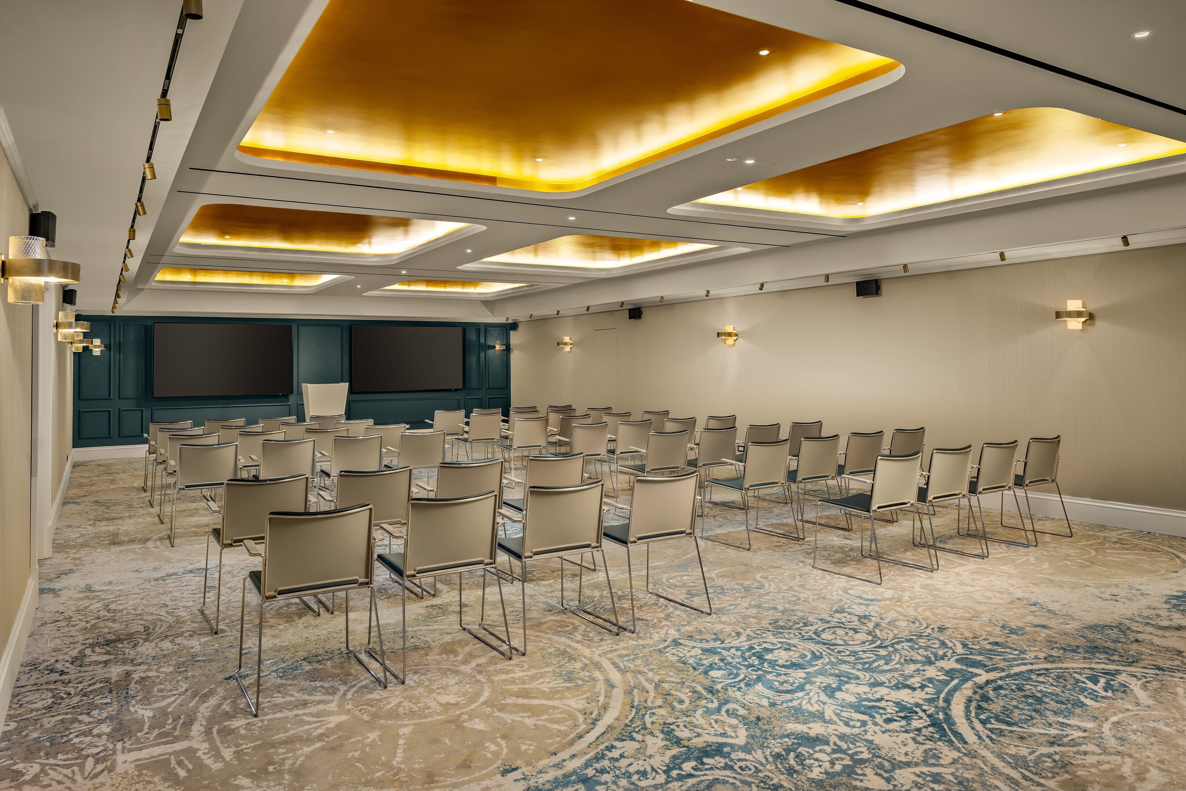 Un salón de conferencias con hileras de sillas, una pantalla grande y una alfombra estampada.