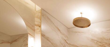 A bathroom with a marble wall, a sink, a mirror, a shower with a gold fixture, and a towel rack.