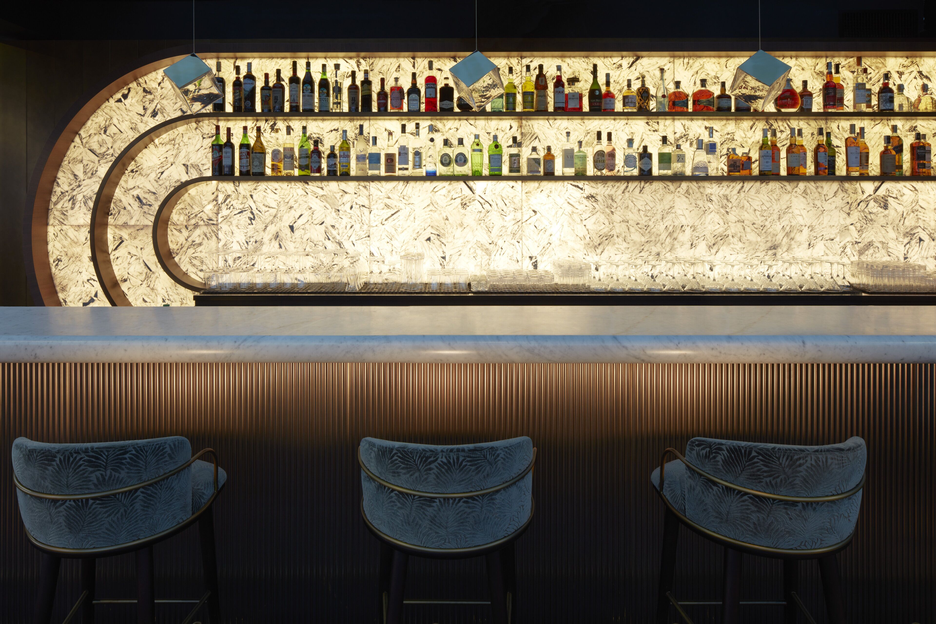 Un bar con mostrador de mármol y varias botellas de licor. Hay tres taburetes tapizados frente al mostrador.