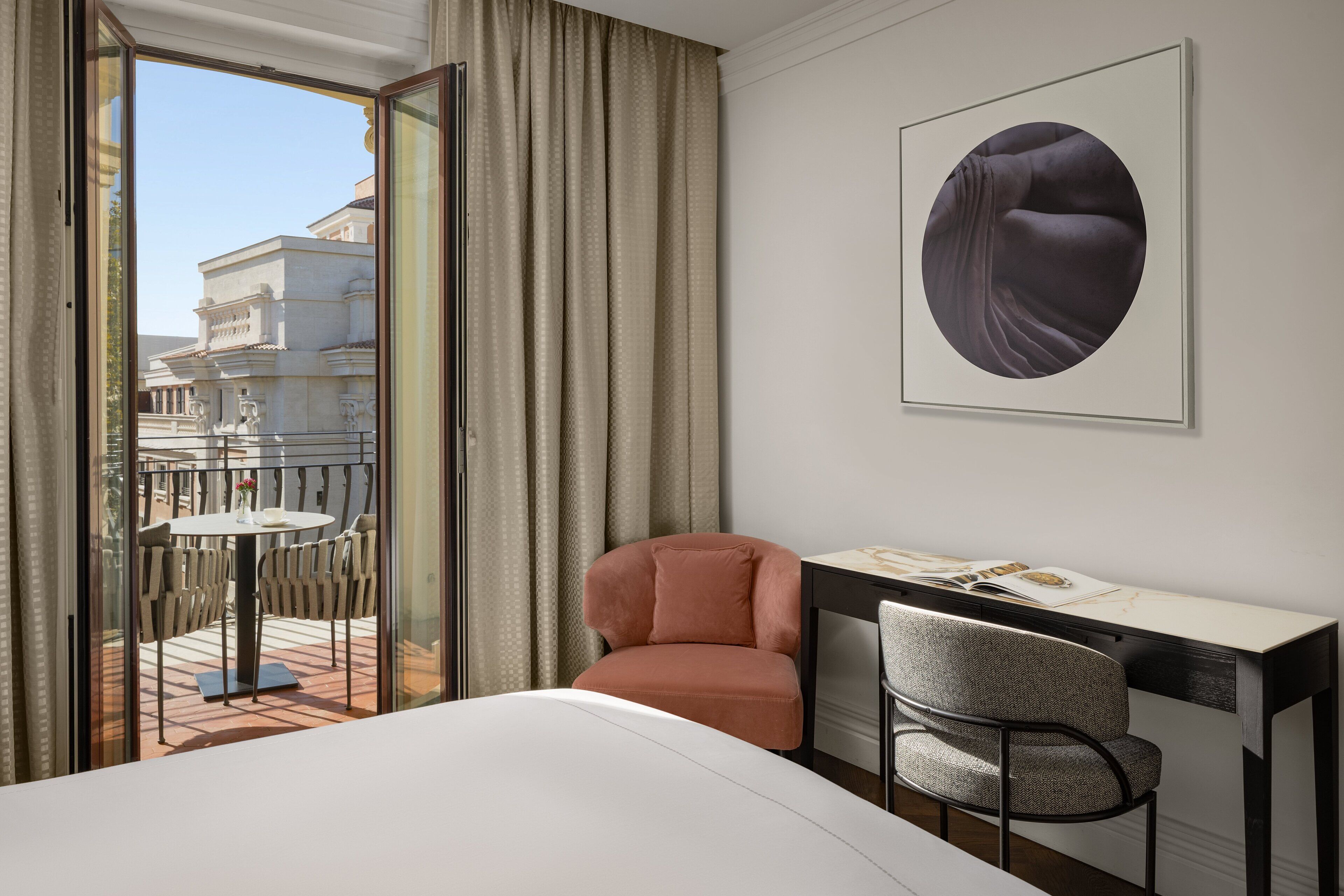 A hotel room with a bed, a desk, a chair, a window with curtains, and a framed artwork on the wall.