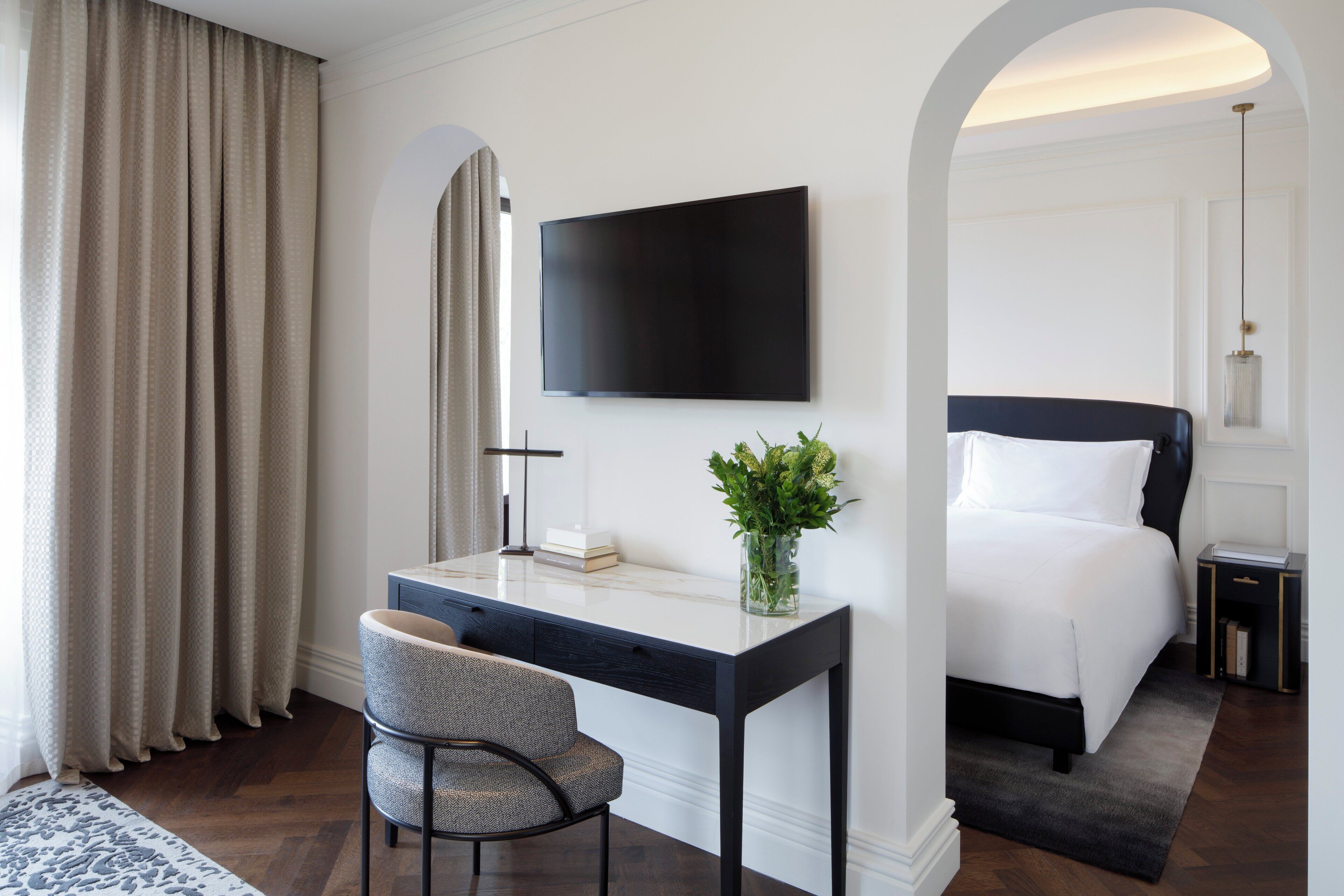 A hotel room with a bed, a desk with a chair, a TV, and a bedside table with a lamp.