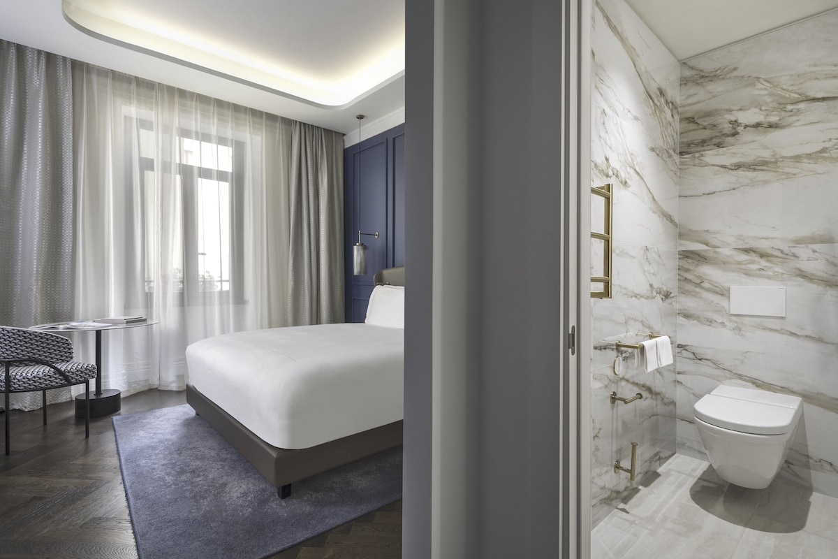 Una habitación de hotel moderna con una cama grande, un escritorio pequeño, una silla y un baño con pared de mármol y un inodoro.