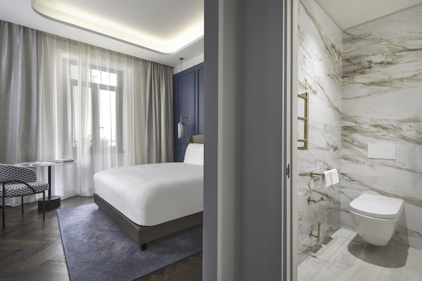 A modern hotel room with a large bed, a small desk, a chair, a bathroom with a marble wall, and a toilet.
