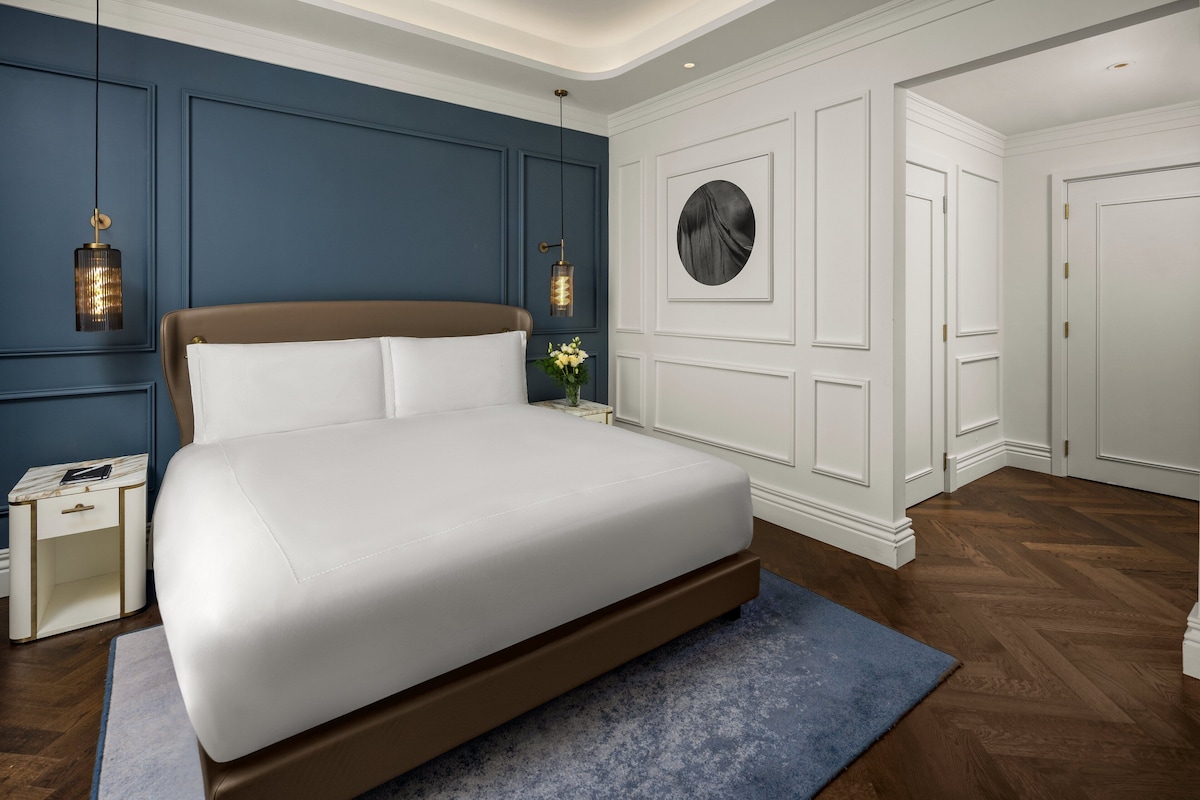 Un dormitorio moderno con una cama grande, mesitas de noche blancas, una pared de acento azul y un espejo redondo.