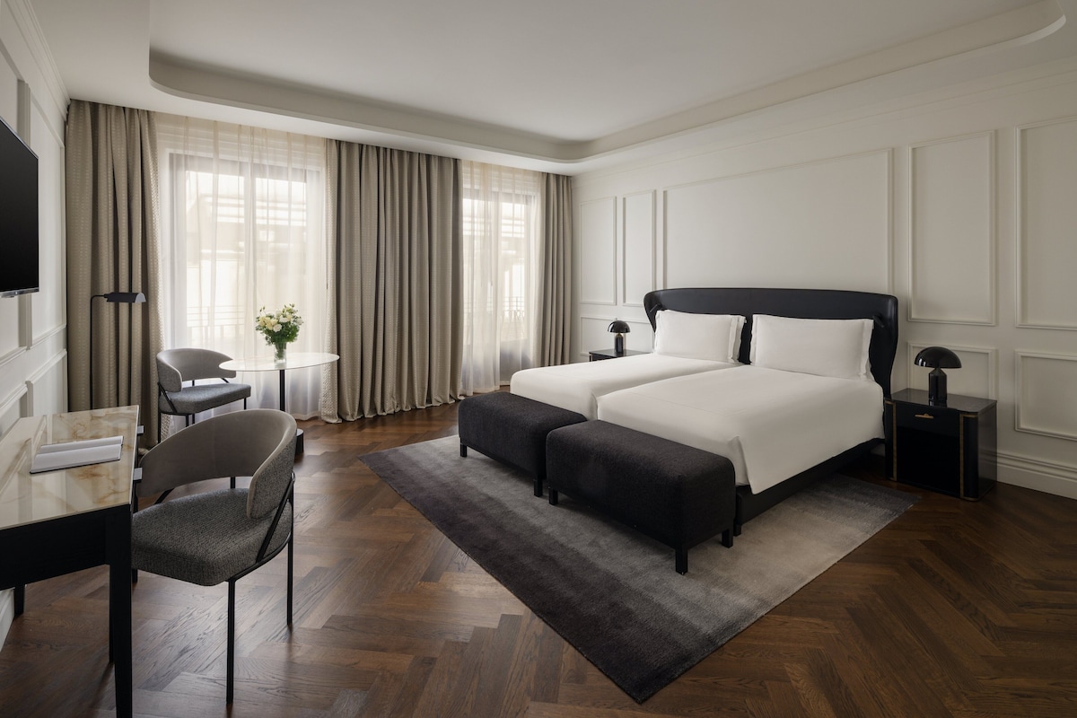 Una habitación de hotel con una cama grande, dos mesitas de noche, un escritorio con silla, una pequeña mesa redonda con otra silla y un ventanal con cortinas.