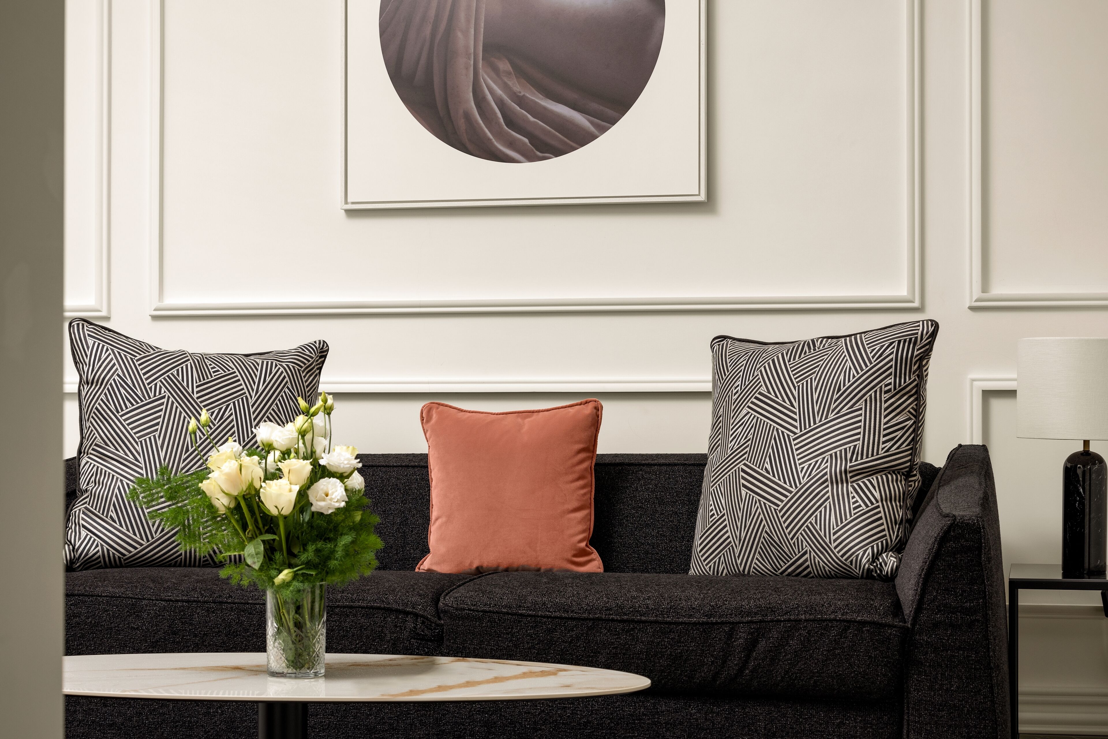 A modern living room with a dark sofa, patterned pillows, a vase with white flowers, and a minimalist artwork on the wall.