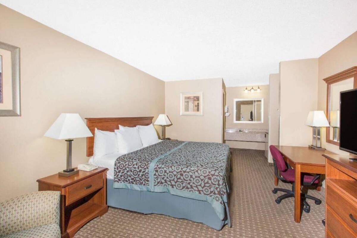 A hotel room with a bed, bedside tables, a desk, a chair, a television, a mirror, and a bathroom visible through an open door.