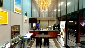 Un hall d’hôtel comprenant un comptoir d’accueil, un lustre, une machine à café et un panneau affichant le menu.