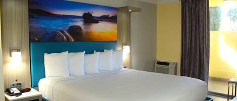 A hotel room with a large bed, a nightstand with a phone and a clock, a painting of a sunset over water on the wall, and a window with curtains.