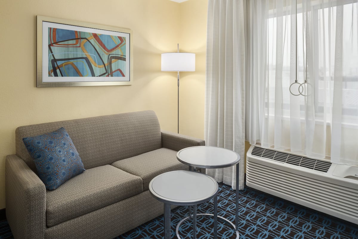 A hotel room with a sofa, two small round tables, a floor lamp, a large window with sheer curtains, and a wall-mounted air conditioning unit.