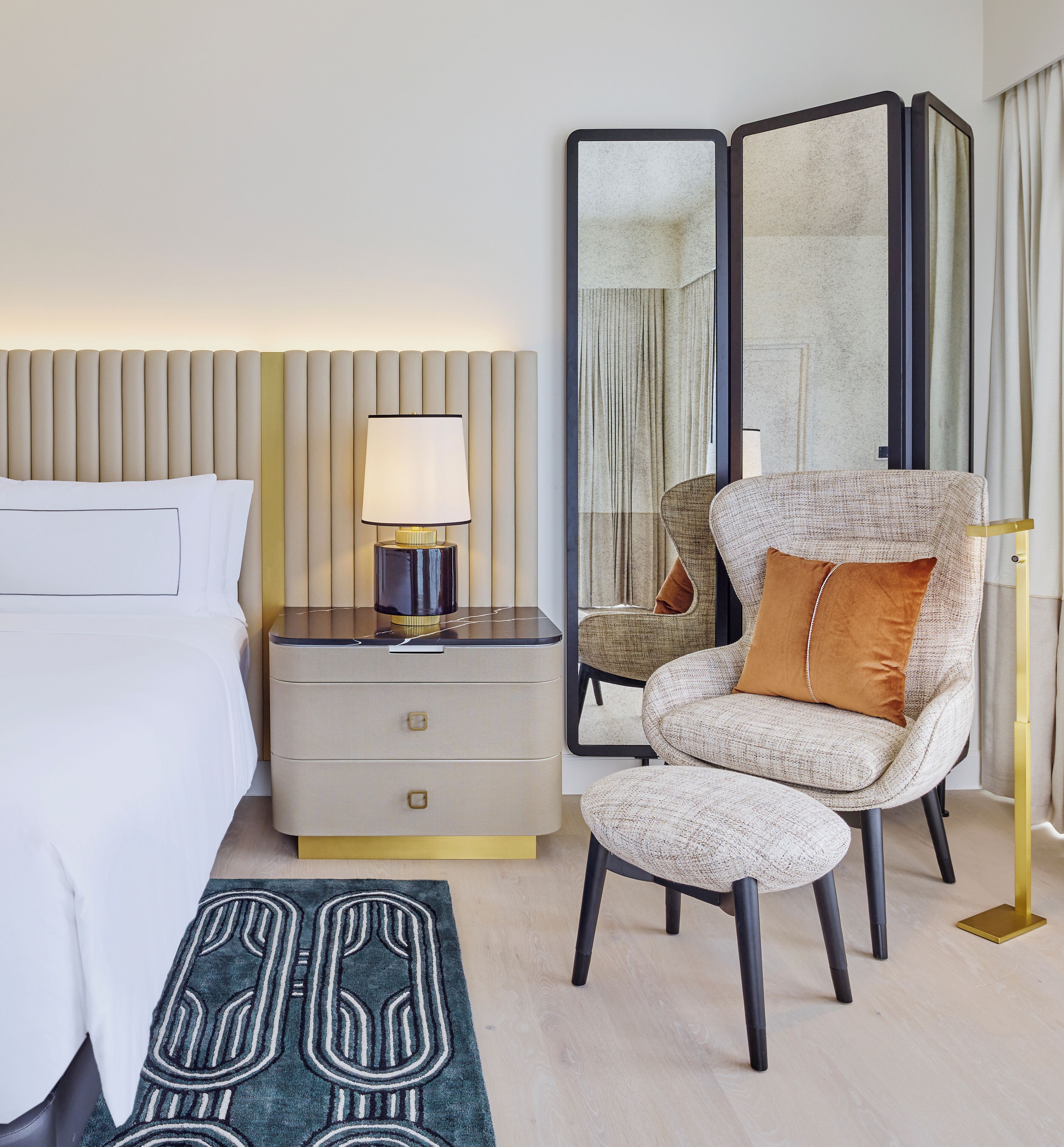 A modern hotel room with a bed, a nightstand with a lamp, a chair with a cushion, a mirror, and a patterned rug.
