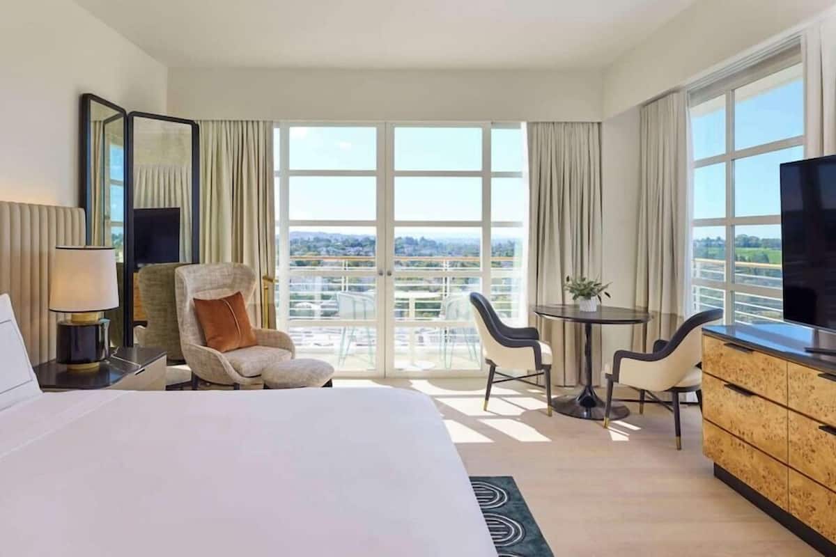 A hotel room with a bed, a chair, a desk, a TV, and large windows offering a city view.