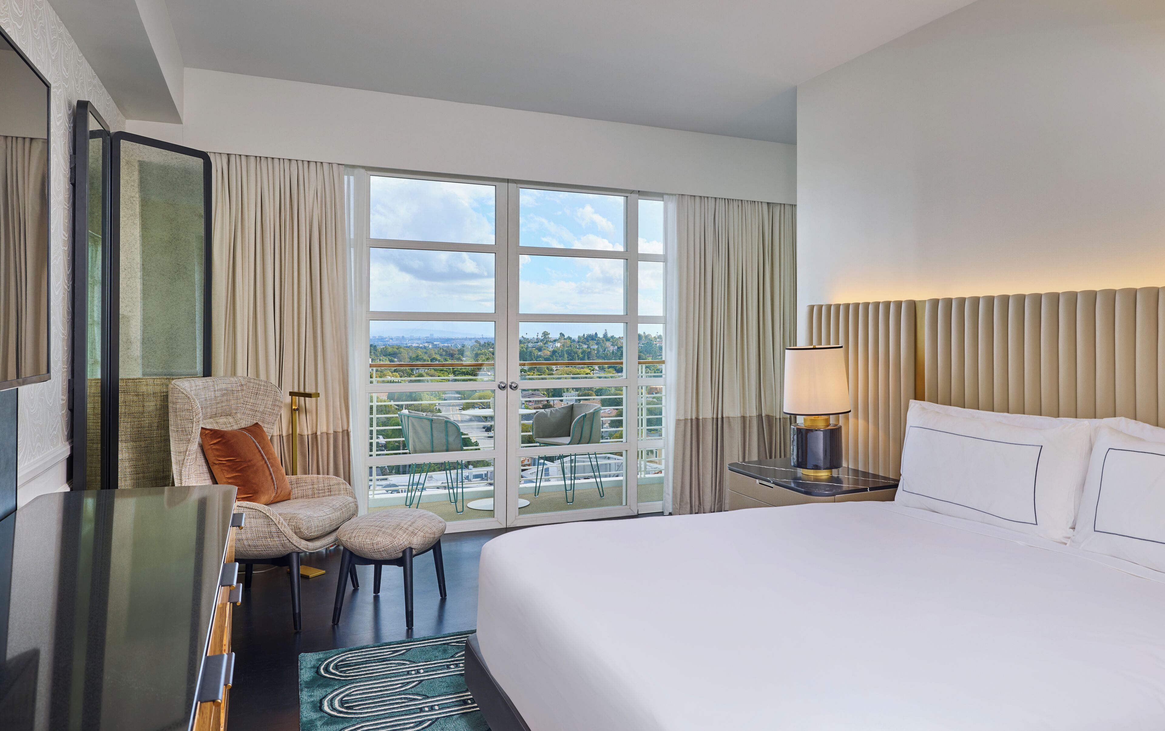 Quarto de hotel com uma cama grande, uma escrivaninha com luminária, uma cadeira e vista da cidade através de janelas do chão ao teto.