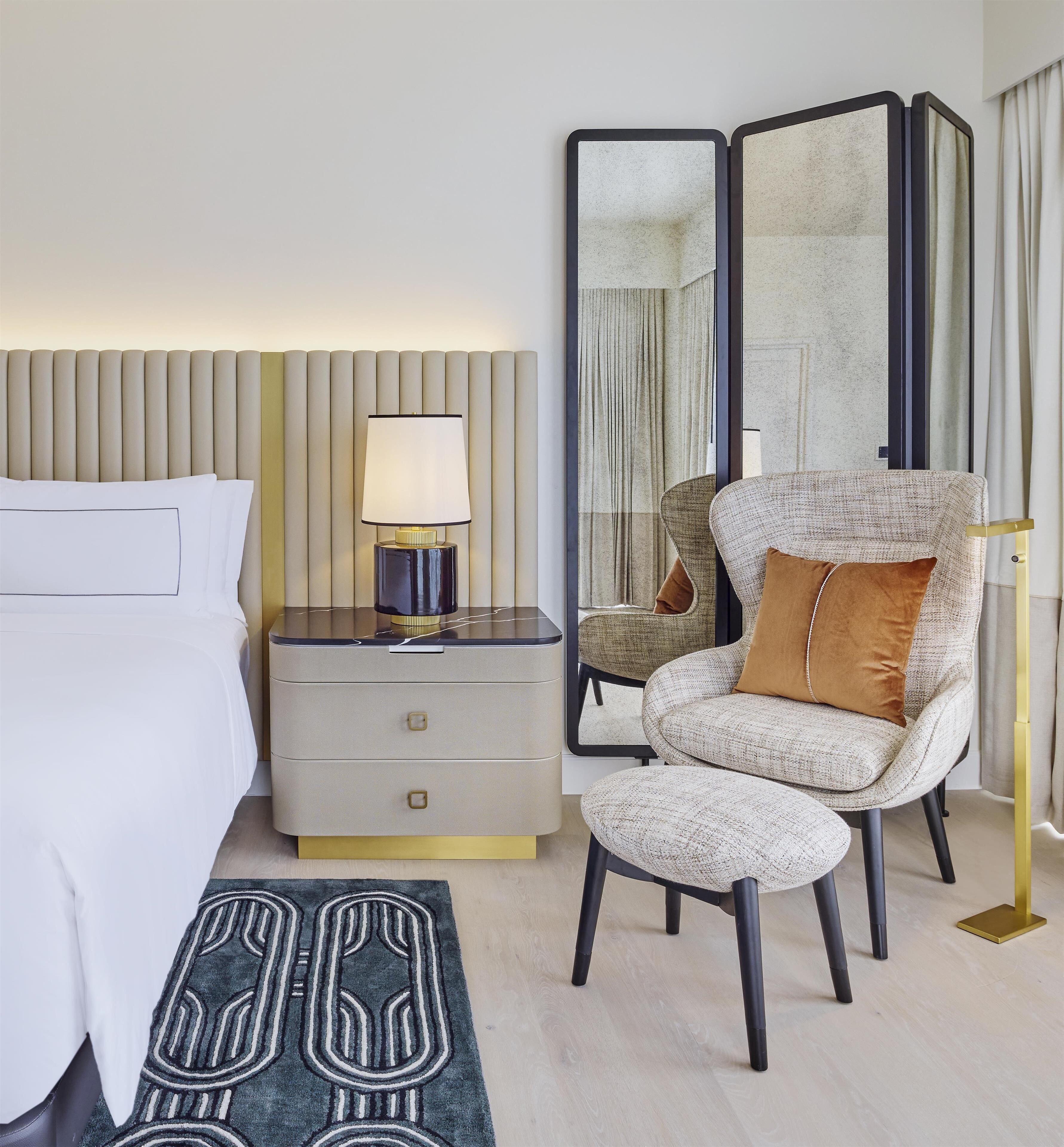 Habitación de hotel con cama, mesita de noche con lámpara, silla con cojín, espejo y alfombra estampada.