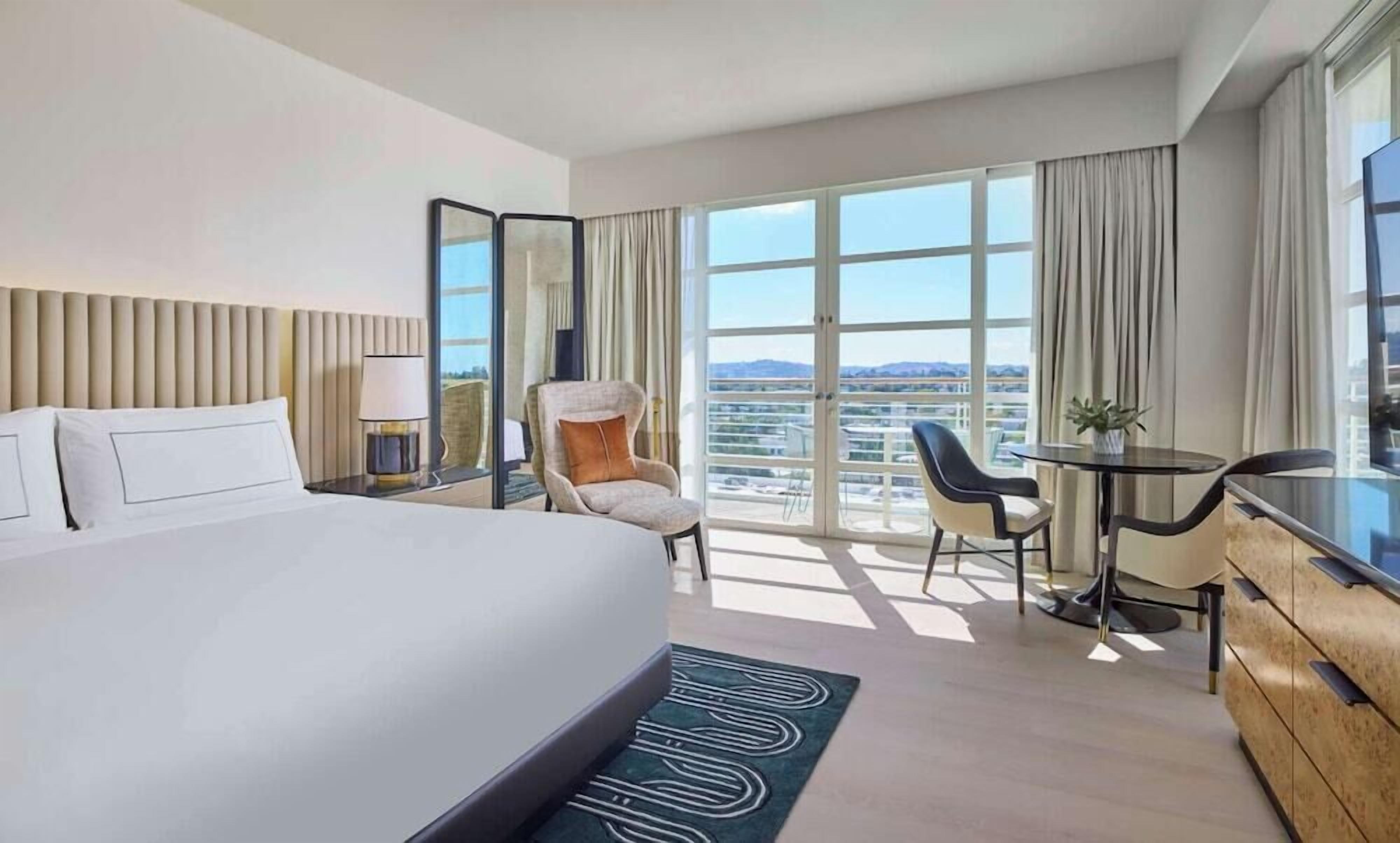Habitació d'hotel amb un llit, una cadira, un escriptori i un finestral gran que ofereix vistes a la ciutat.