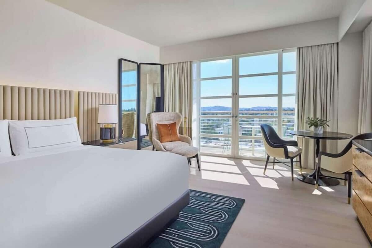 A hotel room with a bed, a chair, a desk, and a large window offering a city view.