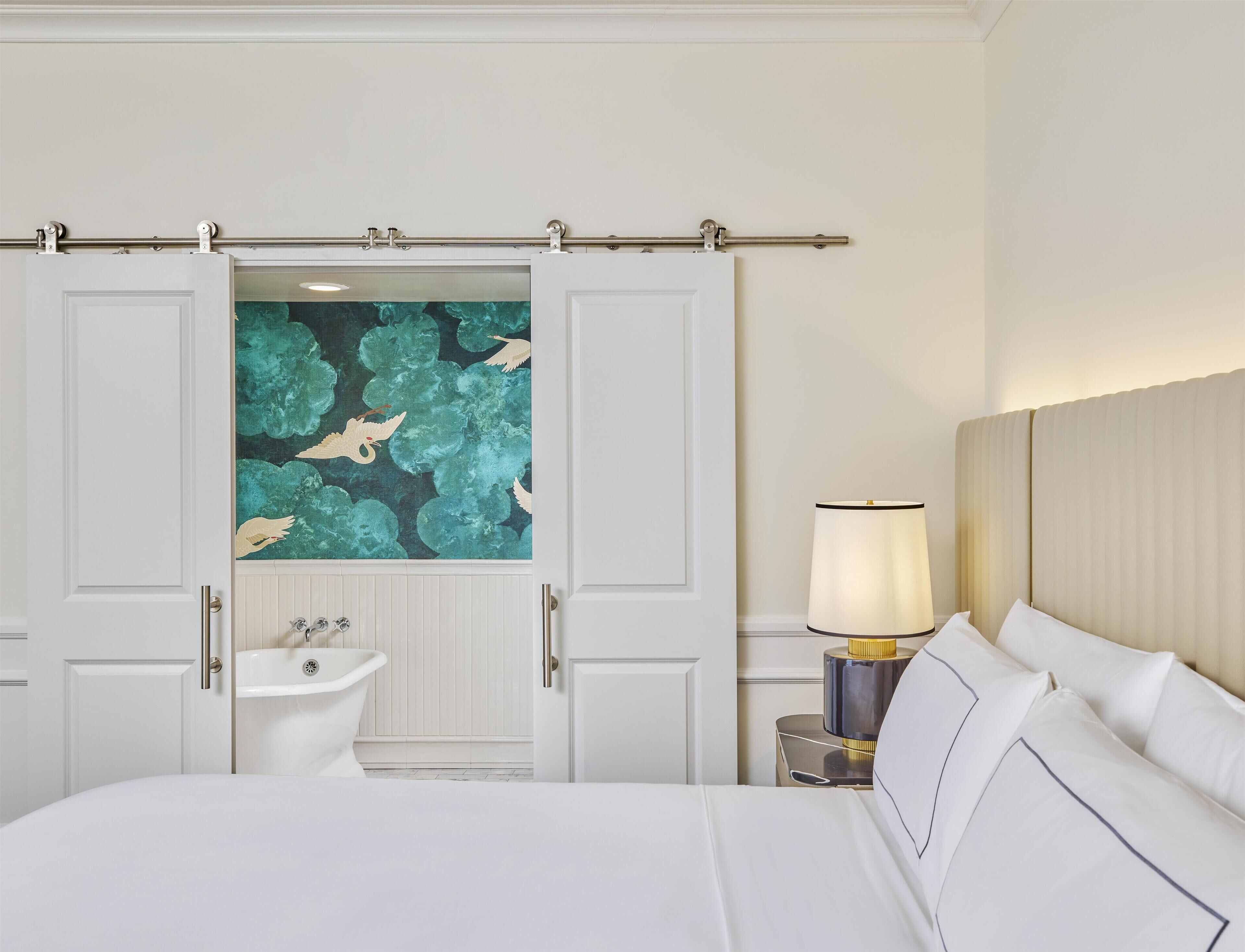 A hotel room with a bed, a nightstand with a lamp, a bathroom with a sink and mirror, and a wardrobe with a teal mural.
