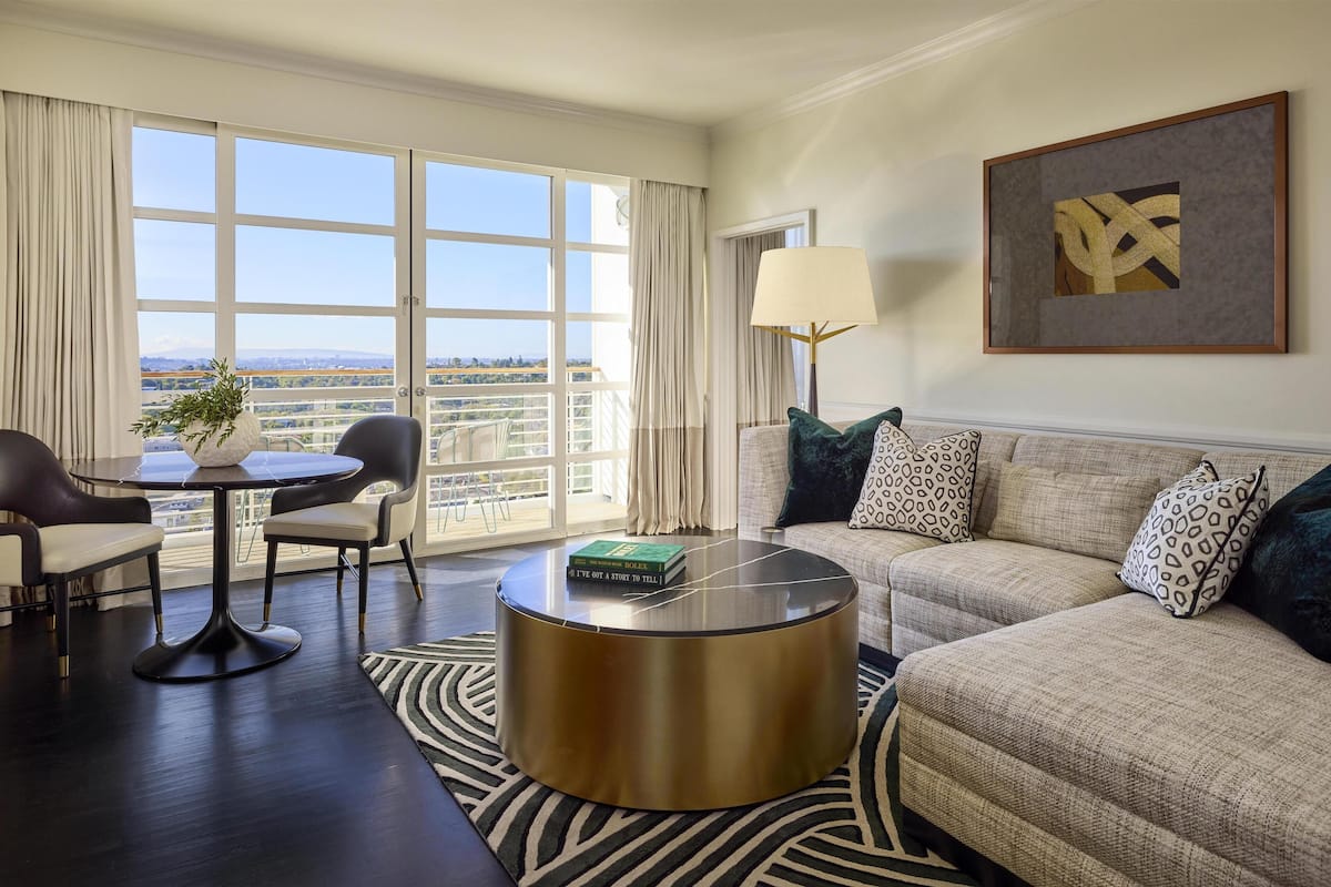 A modern living room with a large window, a round coffee table, a sofa with patterned cushions, and a framed artwork on the wall.