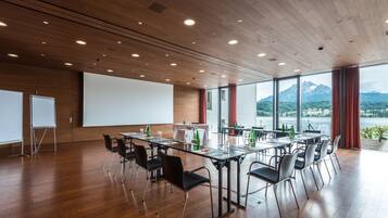 A conference room with a long table, chairs, a projector screen, and a view of a lake and mountains through large windows.