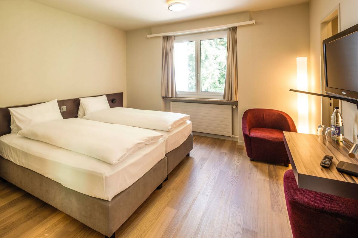 A hotel room with a double bed, a red armchair, a wooden table with a remote control, and a TV.