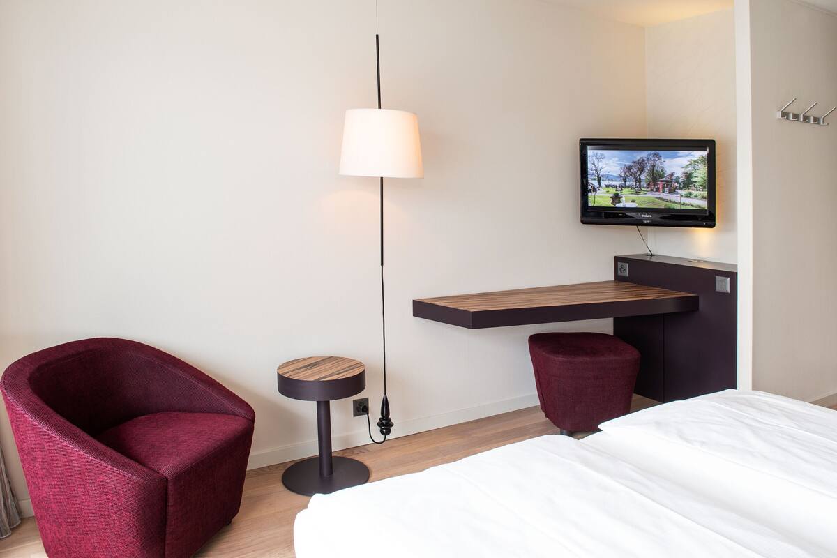 A hotel room with a bed, a red armchair, a small round table, a TV mounted on the wall, and a wooden shelf.