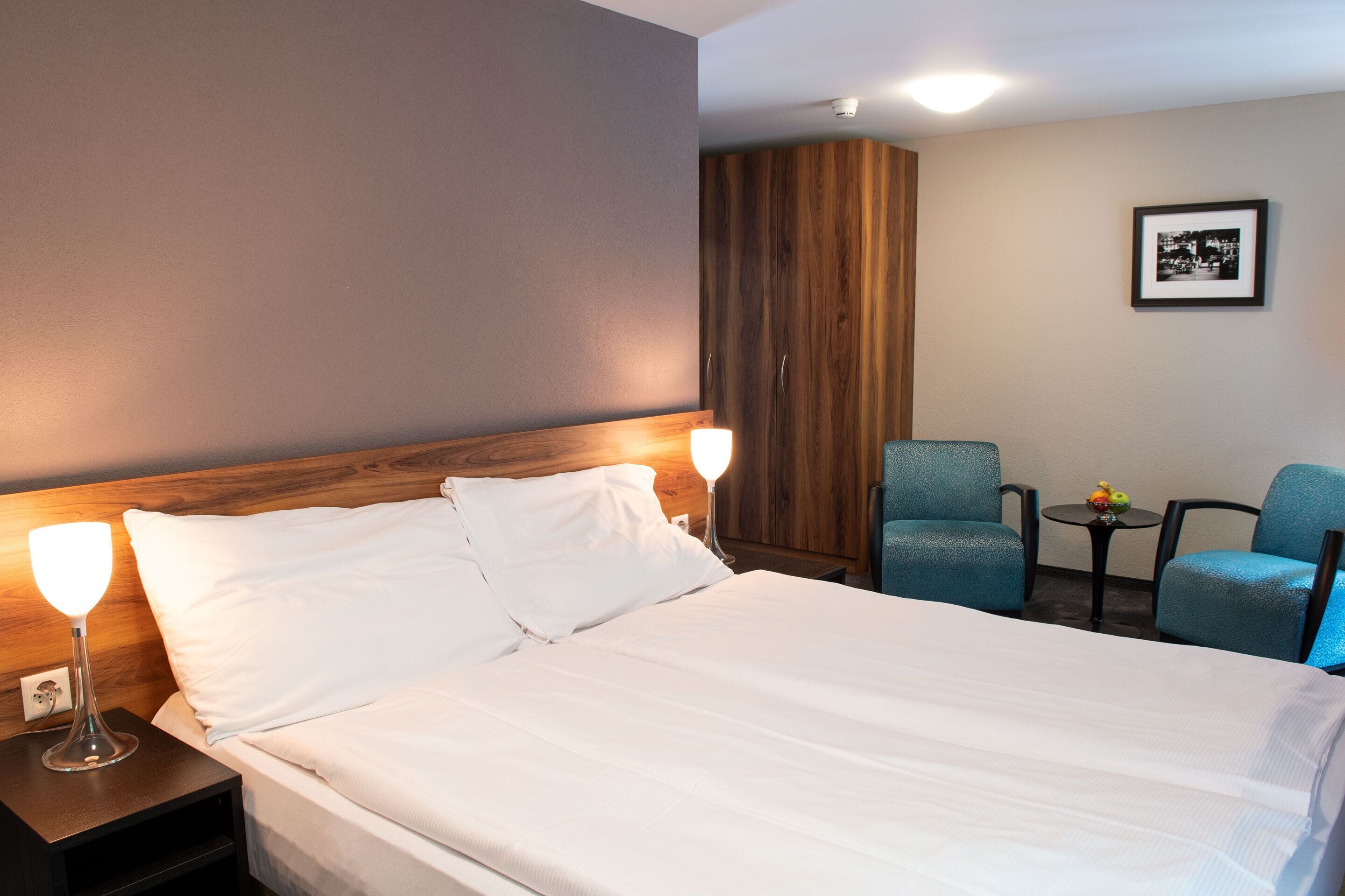 Habitación de hotel con una cama grande, dos lámparas de noche, un armario de madera, una pequeña mesa con fruta y dos sillas azules.