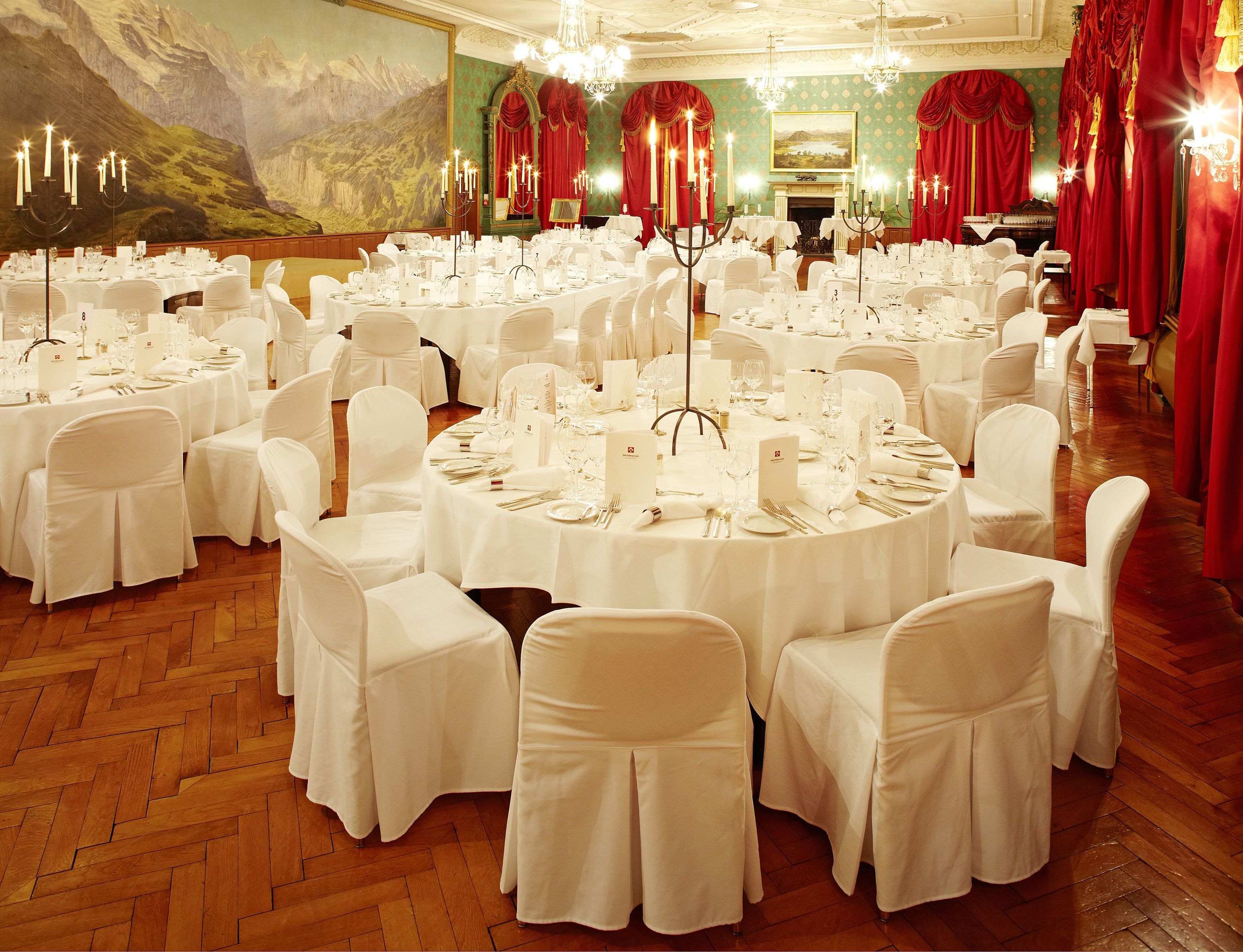 Un salón de banquetes con mesas redondas dispuestas para un evento formal, con un mural grande en la pared y cortinas rojas.