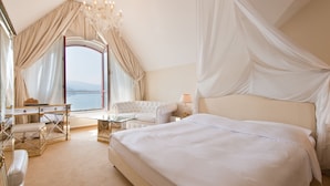 A bedroom with a large bed, a sitting area with a table and chairs, a view of the sea, and a chandelier. - Seeburg Hotel (Lucerne)
