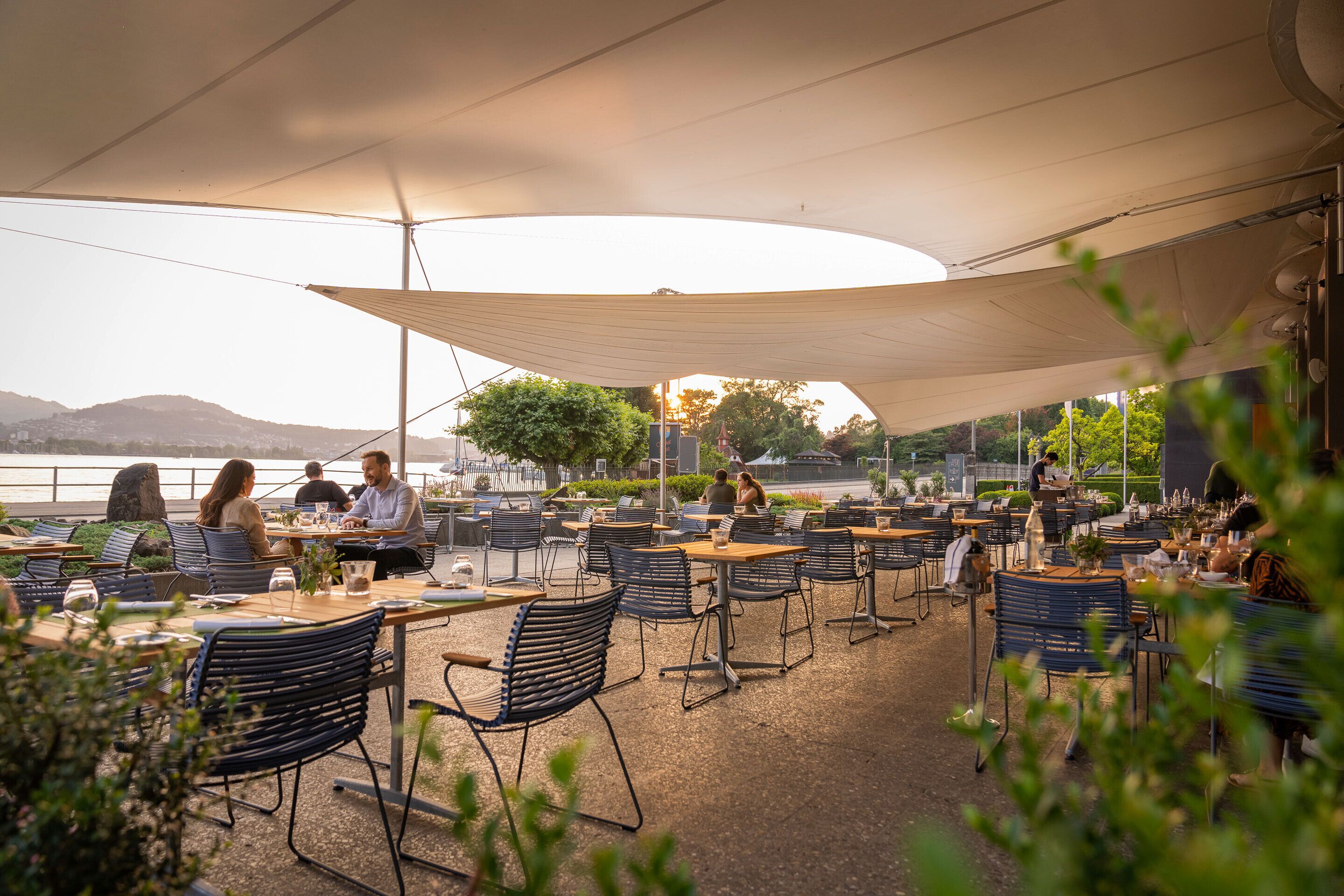Un espacio de comedor al aire libre con mesas y sillas, un toldo sobre la cabeza y vistas a una masa de agua y colinas al fondo.