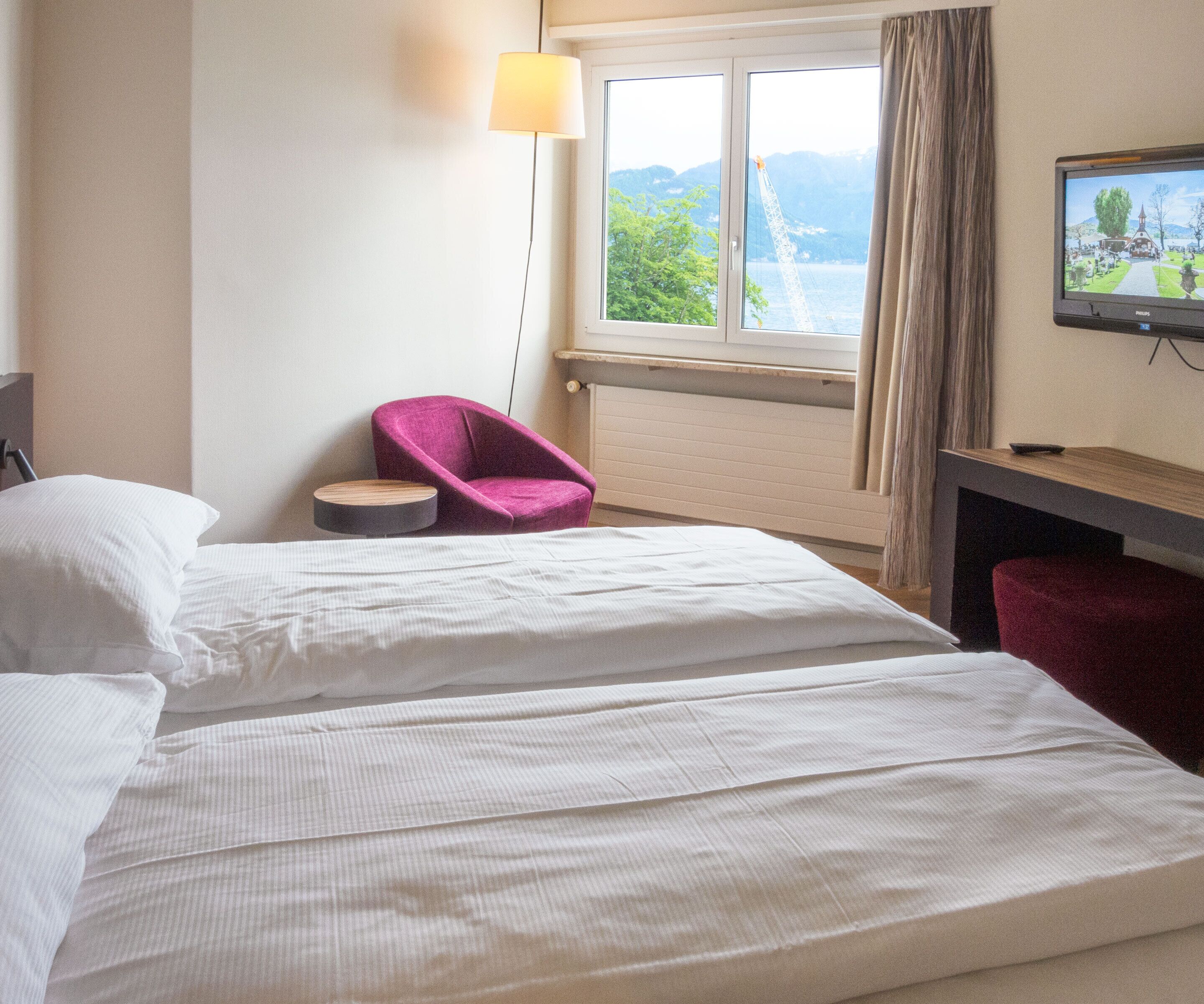 Ein Hotelzimmer mit zwei Betten, einem Schreibtisch mit Fernseher, einem Sessel und einem Fenster mit Blick auf eine Brücke und Grünflächen.
