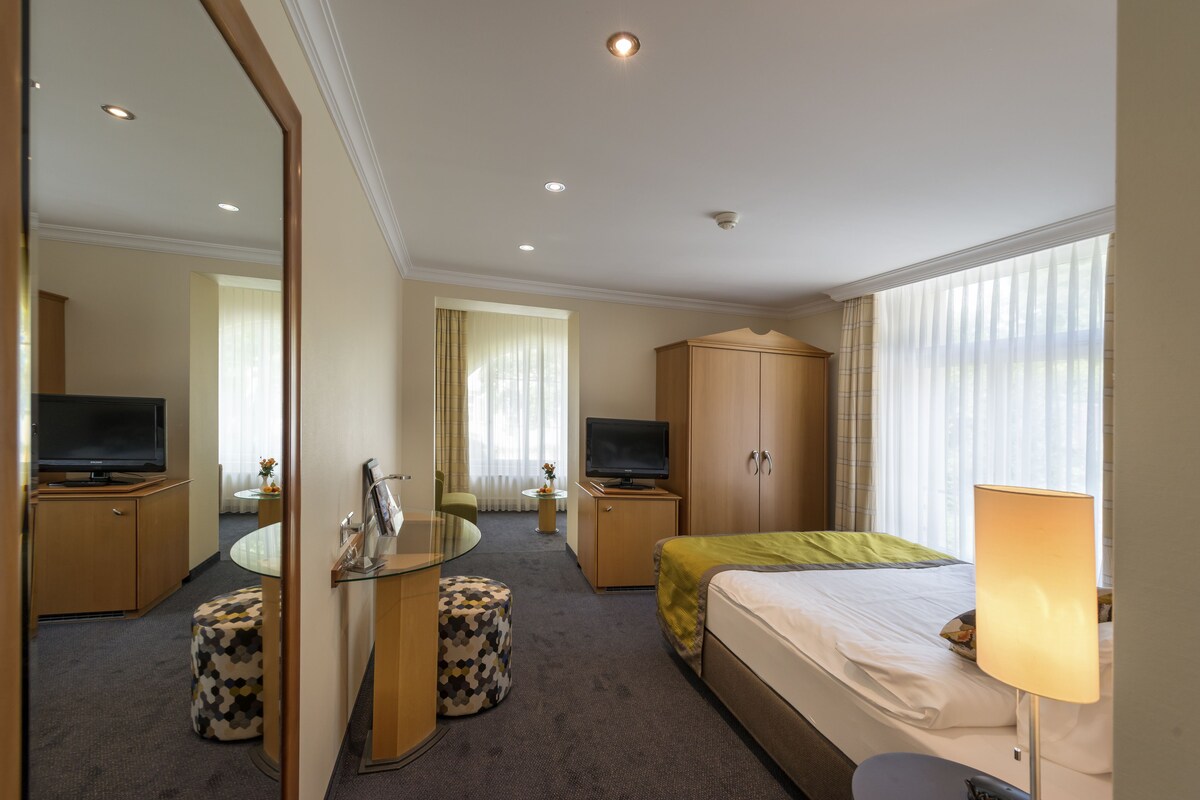 A hotel room with a bed, a desk with a flat-screen TV, a wardrobe, and a glass table.