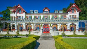 A large, multi-story building with a prominent red entrance, multiple balconies, and a well-maintained garden with hedges and flower beds. - Seeburg Hotel (Lucerne)