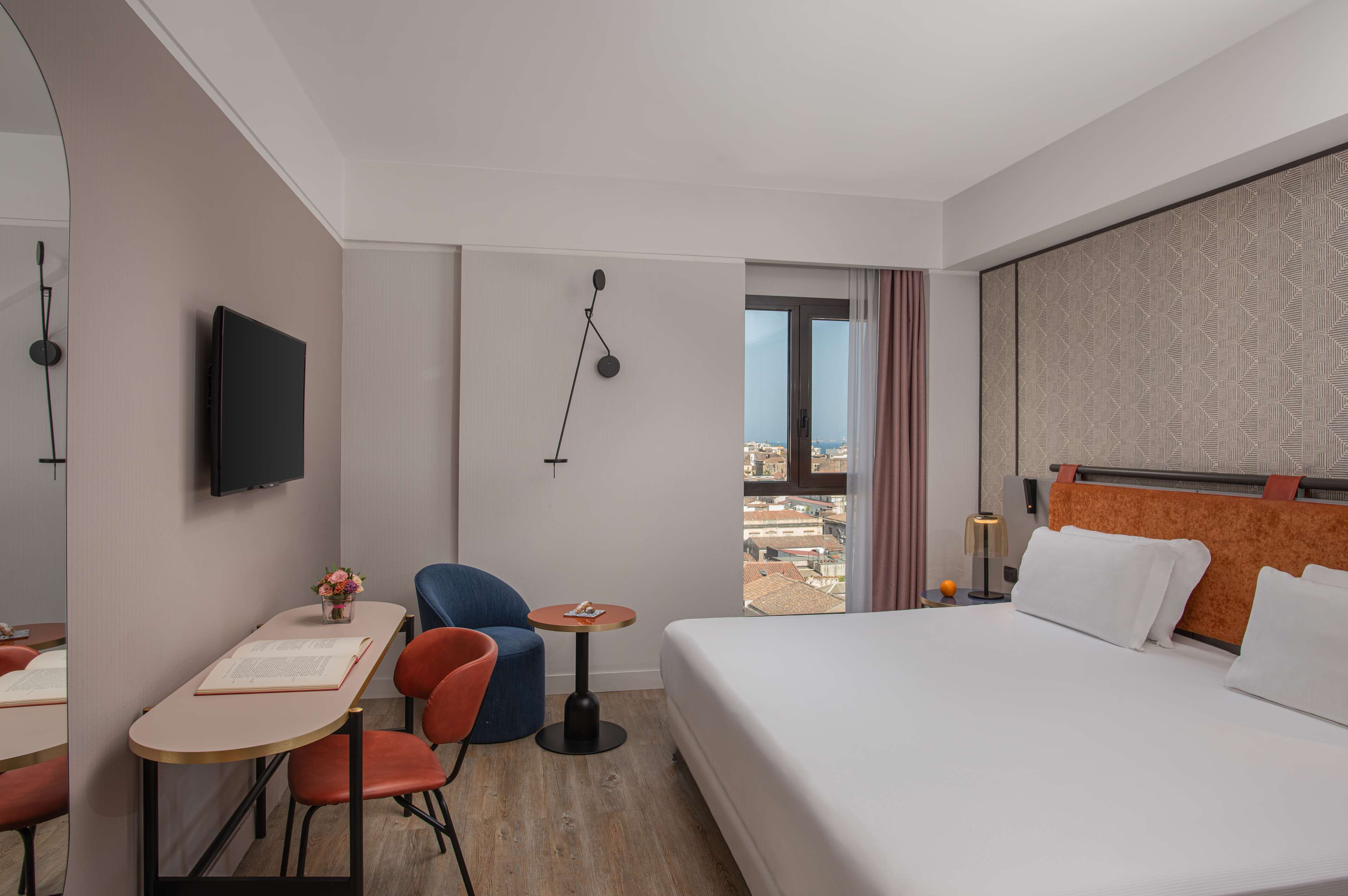 A hotel room with a bed, a desk with a chair, a small table, a TV, and a view of the city through a window.