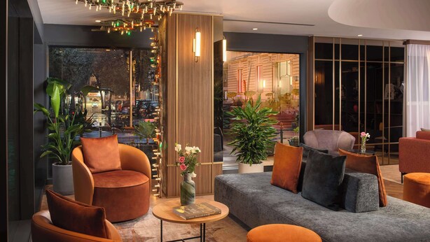 A modern hotel lobby with a seating area featuring orange and grey sofas, a wooden accent wall, and a large window offering a view of the street outside. - NH Catania Centro (Catania)