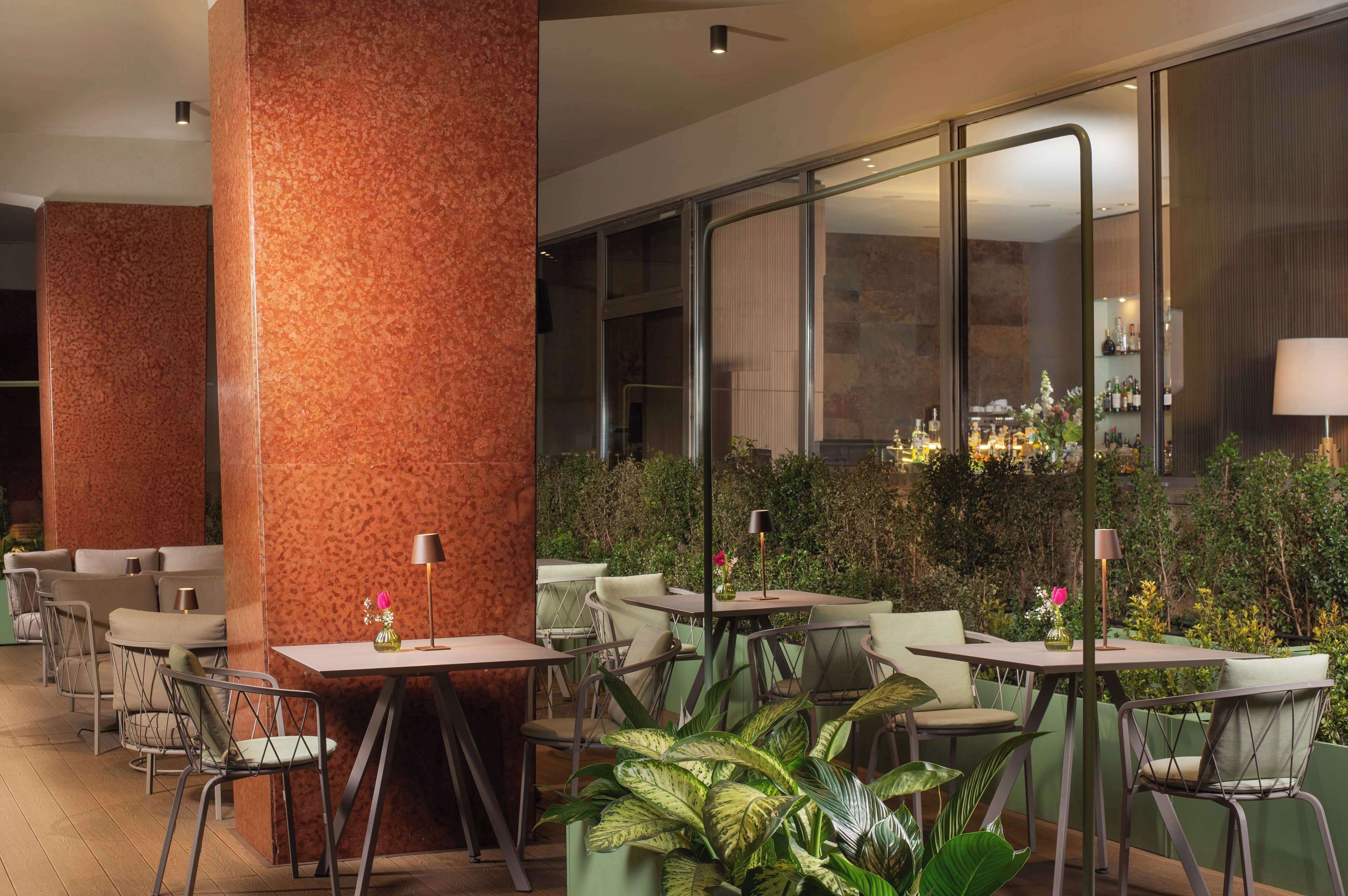 An outdoor dining area with tables and chairs, surrounded by greenery and a glass wall.