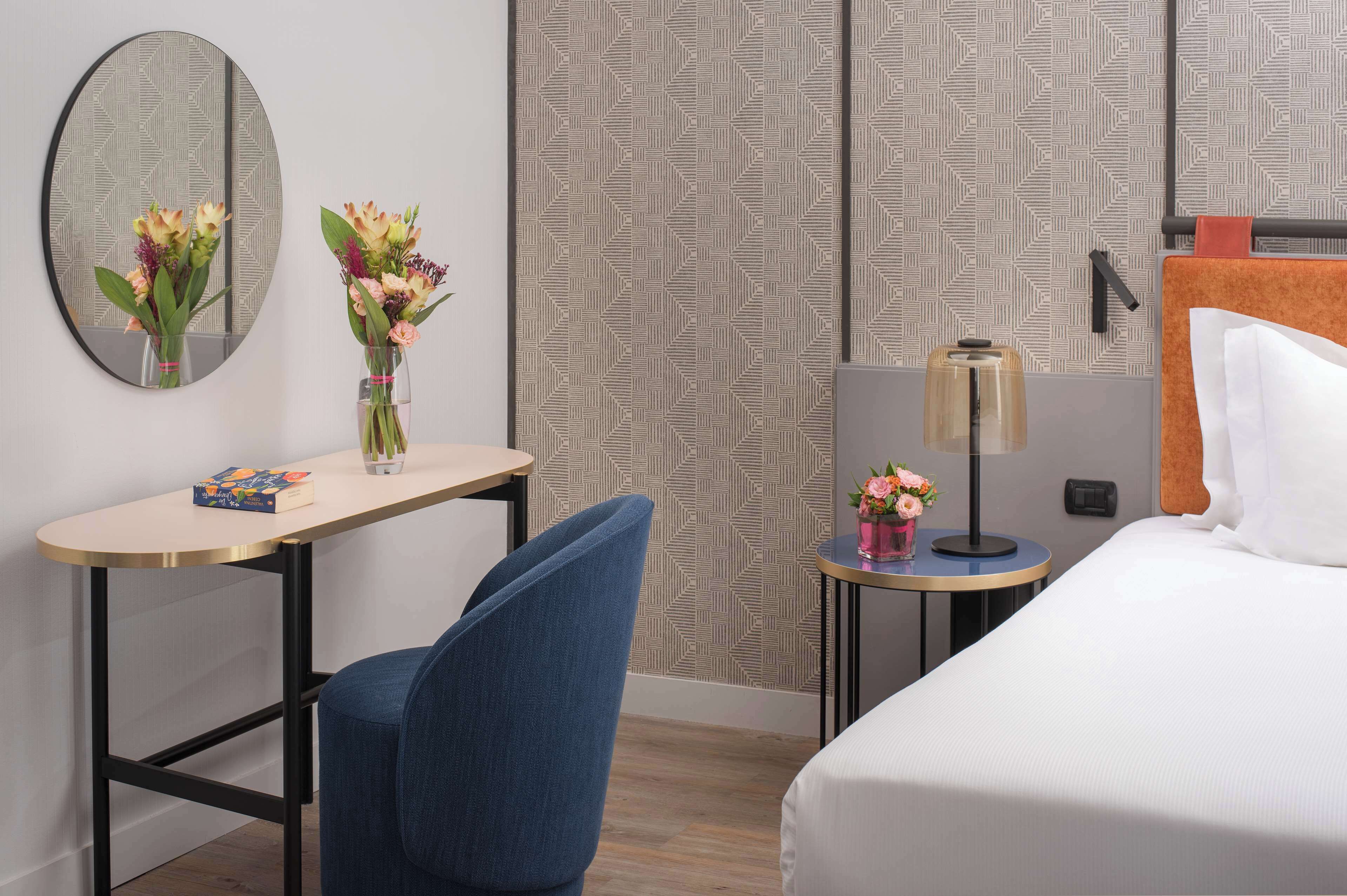 A hotel room with a bed, a desk with a chair, a round mirror, and two vases with flowers.