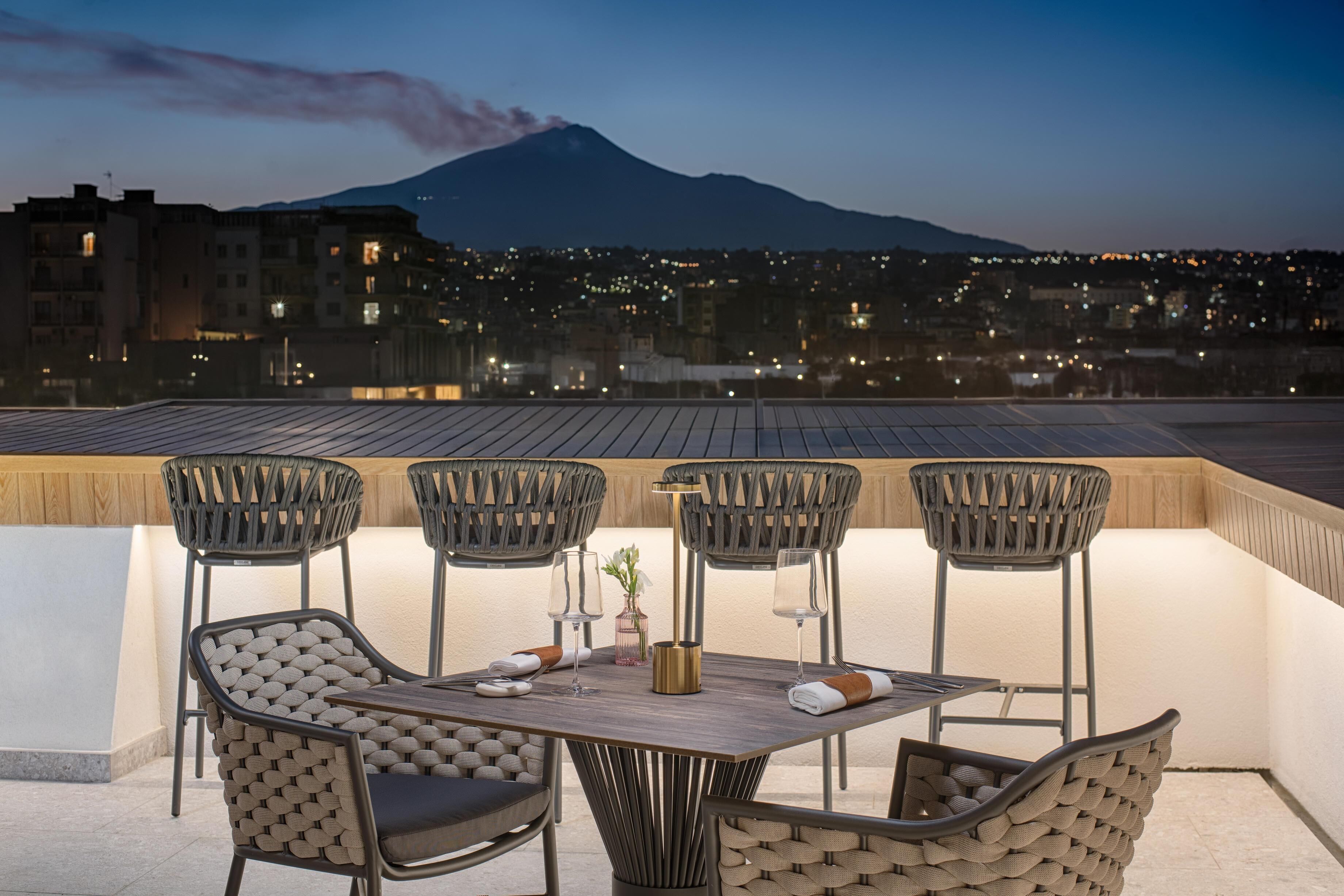 A rooftop terrace with a table and chairs, a view of a mountain, and city lights in the background.