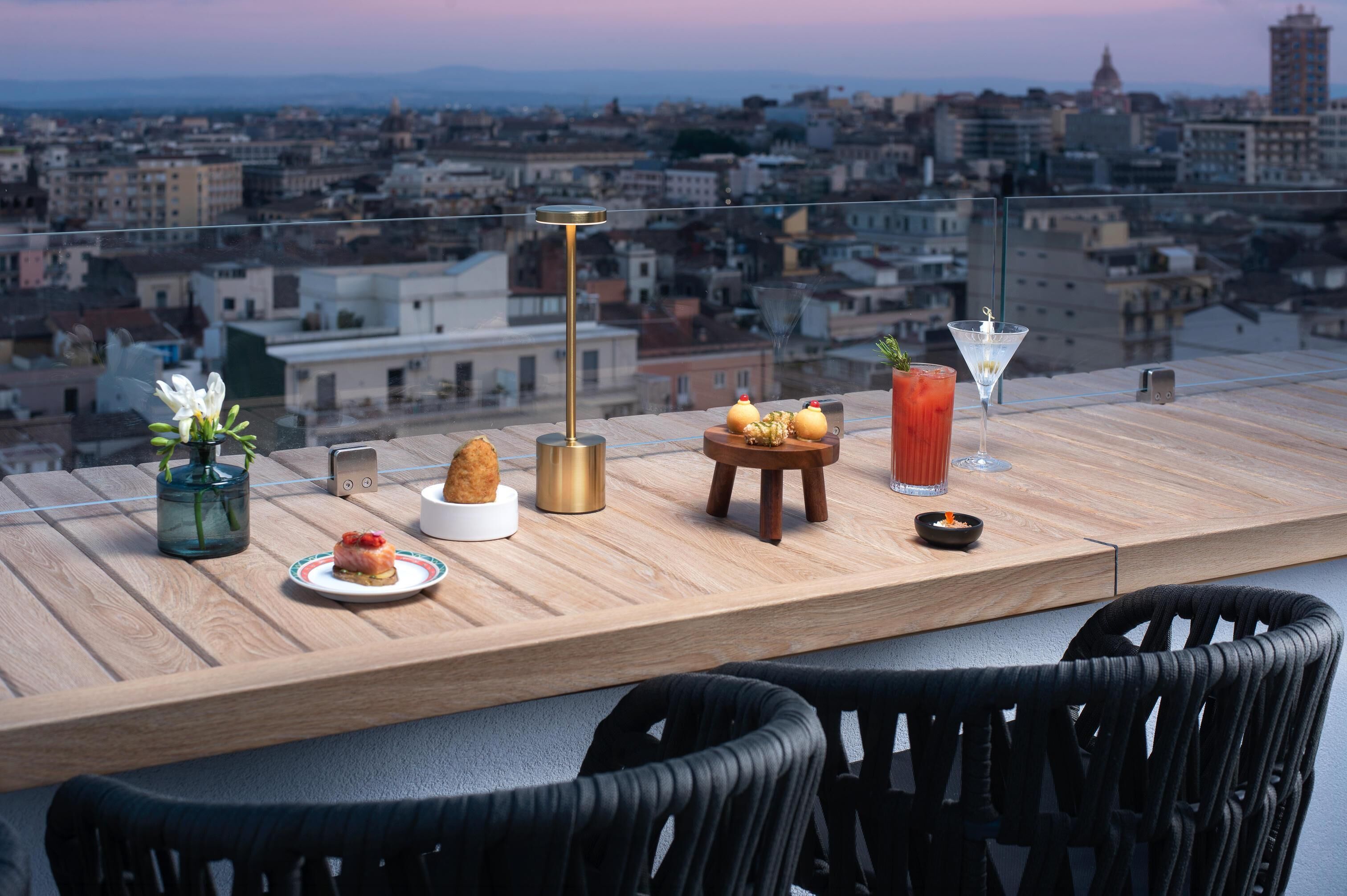 Eine Dachterrasse mit Blick auf die Stadt, mit einem Martini-Glas, einem kleinen Tisch mit Speisen, einer Vase mit Blumen und einer hohen Lampe.