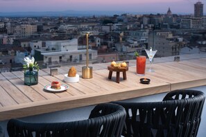 A rooftop bar with a city view, featuring a martini glass, a small table with a dish, a vase with flowers, and a tall lamp. - NH Catania Centro (Catania)