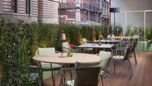 An outdoor patio with round tables and chairs, surrounded by greenery and a building facade in the background. - NH Catania Centro (Catania)