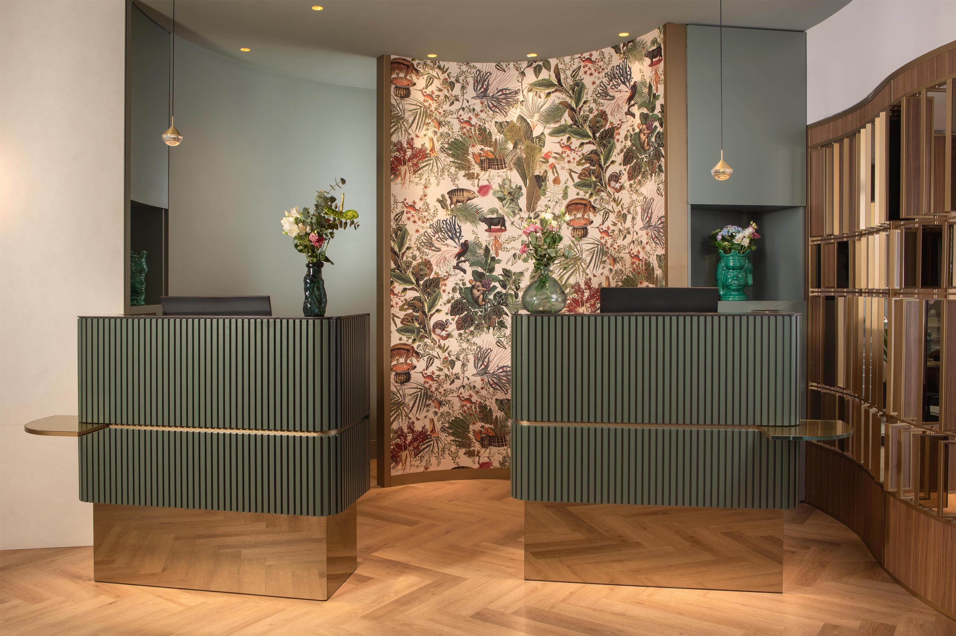 Een receptiezone met twee groene, ribbelachtige balies, een wand met bloemenpatroon en een houten kast met glazen deuren.